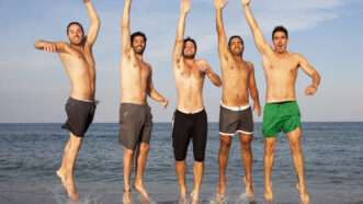 Five men in swim trunks on the beach, jumping into the air with a hand raised. | Adamgregor | Dreamstime.com