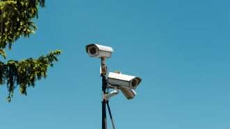 Three security cameras mounted on a pole, facing different directions, against blue sky with a tree branch in the corner | Photo by Michał Jakubowski on Unsplash