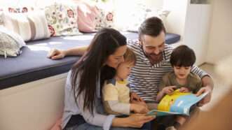 A family with father, mother, and two children sit on the floor reading a book together. | Monkey Business Images | Dreamstime.com