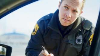 A young male Caucasian police officer, in uniform, leans down to look into a motorist's window as he writes a ticket. | Yuri Arcurs | Dreamstime.com