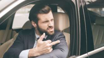 A young bearded man in a suit sits in the driver's seat of a car. He is raising his middle finger at someone not pictured, with a snide look on his face. | Sidelnikov | Dreamstime.com