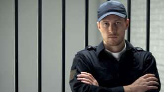 Male prison guard stands with arms folded in front of the bars of a jail cell. | motortion | Dreamstime.com