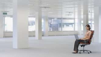 A woman sits alone in a bright, empty office space. She sits in an office chair with a laptop computer in her lap. | Photographerlondon | Dreamstime.com