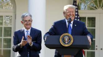 Donald Trump laughs, stands in front of a podium with the presidential seal on it, while Jerome Powell claps behind him and part of the White House's exterior is in the background. | Polaris/Newscom