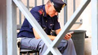 An Italian police officer guarding a jail cell sits in a chair looking at his smartphone. | Angelo Cordeschi | Dreamstime.com