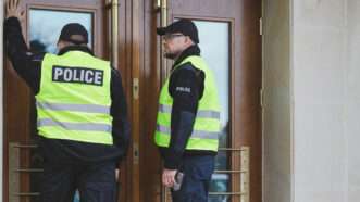 Two police officers in neon "POLICE" vests stand at a building's entrance, in front of closed doors. | Katarzyna Bialasiewicz | Dreamstime.com