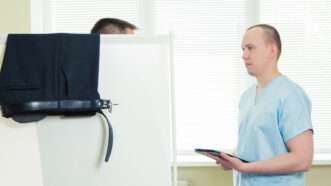 A doctor in blue scrubs speaks to a patient, who is concealed behind a divider, in a doctor's office. | Aleksey Satyrenko | Dreamstime.com