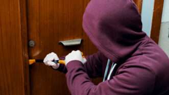 A burglar in a hoodie uses a crowbar to break into a locked door. | F01photo | Dreamstime.com