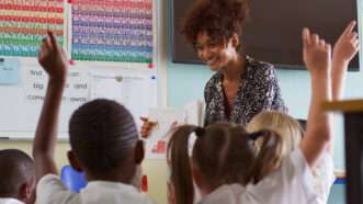 A teacher holds a book in front of her class, which is seated on the floor in the classroom. Many of the students have their hands raised. | ID 343799779 ©  Monkey Business Images | Dreamstime.com