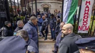 People in law enforcement standing in front of a fence with banners hanging from it, with people in the background, on the other side of the fence, holding up phone cameras. |  Michael Nigro/Sipa USA/Newscom