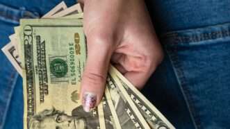 Close-up of a woman's hand behind her back, clutching fanned-out cash in 20s and 50s. | Vlad Ispas | Dreamstime.com