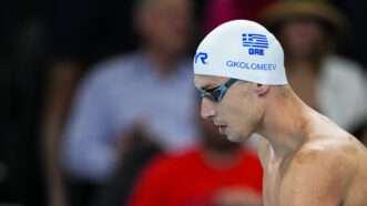 A man in the foreground, wearing swimming goggles and a swimming cap with the Greek flag on it. A few people are in the audience in the background, sitting. | Oscar J. Barroso/ZUMAPRESS/Newscom