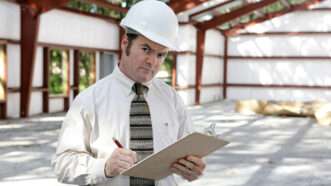 A code inspector in a hard hat stands in a building under construction, holding a clipboard and giving the camera a suspicous look. | Brian T. Young | Dreamstime.com