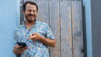 A man in a floral-print shirt laughs while pointing at his cell phone, as if he has just seen something funny. | Jose Maria Ruiz | Dreamstime.com