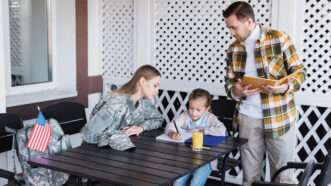 A young girl does homework at an outdoor table, surrounded by her mother—who is wearing combat fatigues—and her father. | Lightfieldstudiosprod | Dreamstime.com