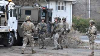A group of law enforcement officers in camouflage and SWAT gear are assembled outside a suburban home, preparing to search for a suspect. | Kyle Mazza/ZUMAPRESS/Newscom