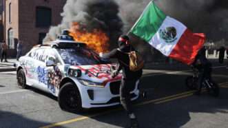 Protester in Los Angeles riots in front of burning car and waving a Mexican flag | Caylo Seals/Sipa USA/Newscom