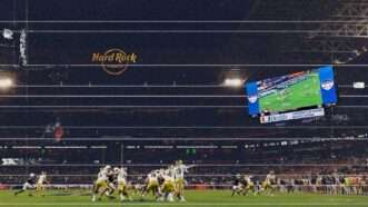 A weather image of an overview of Hard Rock Stadium during a football game, with many Notre Dame football players on the ground in white jerseys and gold helmets, and a large video board in the right side of the stadium. | Illustration: Eddie Marshall | Midjourney. Photo: Joe Robbins/Icon Sportswire 573/Joe Robbins/Icon Sportswire/Newscom