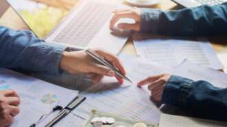 Two people sitting across from each other work on a financial audit, with cash and spreadsheets on the table. | Lovelyday12 | Dreamstime.com