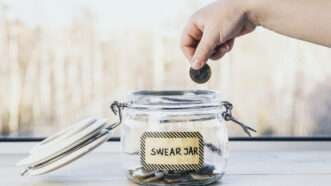 A child's hand places a coin into a clear jar filled with money, labeled "Swear Jar." | Helinloik | Dreamstime.com