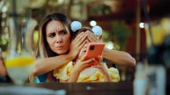 A mother holds hands over her daughter's eyes as the daughter holds a cell phone. | Nicoleta Ionescu | Dreamstime.com