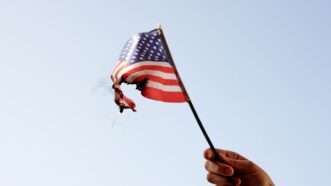 A hand holds a small, singed American flag in the air, as it lightly burns. | Mahbub Hasan | Dreamstime.com