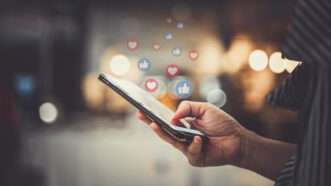A hand holds a smartphone, as heart and thumbs-up emoji reactions waft off the screen. | Kittichai Boonpong | Dreamstime.com