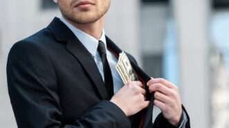 A man in a business suit stuffs cash into his jacket pocket. | Gstockstudio1 | Dreamstime.com