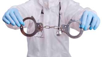 A doctor wearing scrubs and medical gloves holds up a pair of handcuffs. | Andrey Zhuravlev | Dreamstime.com