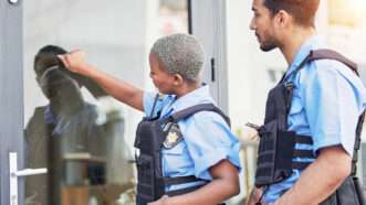 A black female police officer knocks on a residential storm door while a white male officer stands behind her. They both wear uniforms and body armor. | Yuri  Arcurs | Dreamstime.com