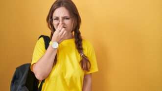 A young woman with a backpack over one shoulder holds her nose, as if she smells something bad. | Aaron Amat | Dreamstime.com