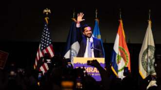 Zohran Mamdani speaks to a crowd behind a podium with his campaign sign attached and with flags in the background | Lev Radin/Sipa USA/Newscom