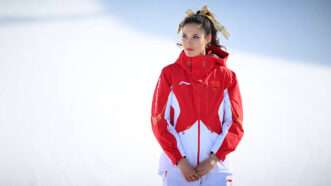 In front of a snowy white background, Eileen Gu stands facing the camera with her hands clasped, wearing a white and red jacket with the Chinese flag on it. | IMAGO/REAU ALEXIS/IMAGO/PRESSE SPORTS/Newscom