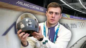 In the background is the curve of a sliding track with the Milano Cortina 2026 Olympics branding, in front of the background is a separate image of Vladyslav Heraskevych holding up a gray helmet with several faces on it. | Illustration: Kyodonews/ZUMAPRESS/Imagine China/Newscom