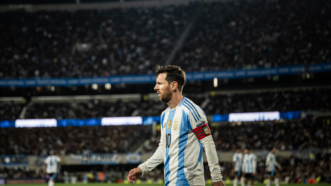 Lionel Messi walking on a soccer field, with several other Argentina players in the background, and three full tiers of stadium seating behind them. | Manuel Cortina / SOPA Images/Sipa USA/Newscom