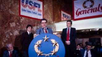 New Jersey Generals coach speaks at a lectern during a press conference, flanked by team owner Donald Trump and quarterback Doug Flutie. | Photo: Bernard Gotfryd/Wikimedia Commons