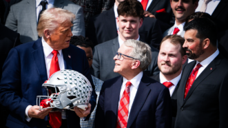 President Donald Trump holds an Ohio State football helmet, while Ohio Governor Mike DeWine and head coach Ryan Day all look at him. Various members of the football team wearing suits are in the background. | Tom Williams/CQ Roll Call/Newscom