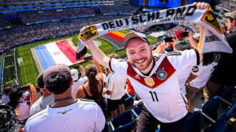 A man in a 2014 Germany soccer jersey holds up a scarf that says DEUTSCHLAND on it, standing in the stands above a soccer field with the Paraguayan and German flags on it. | Jason Russell