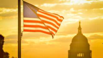 The American flag flaps in the breeze at dusk. The dome of the U.S. Capitol is visible in the distance. | Orhan Çam/Dreamstime