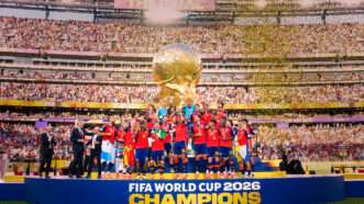 The Spanish soccer team celebrates on a stage in front of a stadium crowd and a large, golden World Cup trophy in the background. | Photo: EyePress/Newscom