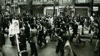 A black-and-white photo of people marching in a street, all toward the left side of the photo frame. A few of them hold signs on posts. | Wikimedia Commons