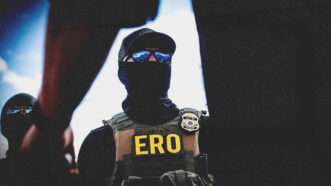 A masked agent with Immigration and Customs Enforcement's (ICE) Enforcement and Removal Operations (ERO) badging on his body armor. He wears sunglasses and a baseball cap, in addition to a gator-style face mask. | Kelly Kimball/ZUMAPRESS/Newscom