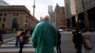 An elderly man with white hair stands on a sidewalk in a green hospital gown with the CN Tower in the background. | Illustration: Adani Samat/Midjourney. Photo: Nadtochiy/Dreamstime