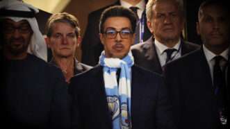 People in suits stand and look out, in the center of the frame is a man in glasses with a light blue and white Manchester City scarf tied around his neck. | Shaun Brooks/Action Plus/Newscom