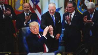 President Donald Trump holds Sen. Dan Sullivan's (R–Alaska) hand in the Oval Office, surrounded by other people. | Illustration: YURI GRIPAS/UPI/Newscom