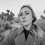 A woman with blonde hair wearing a green jacket in a selfie in front of Joshua trees. | Liz Wolfe