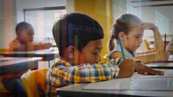 Young students sitting at desks with pencils in their hands. A very translucent layer of graphs and spreadsheets is laid over the photo of the students. | Wave Break Media Ltd/Dreamstime