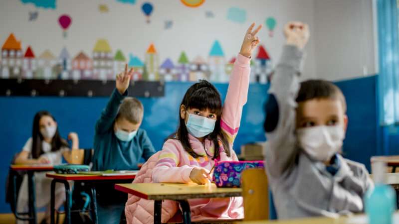 Masked children raise hands in a classroom