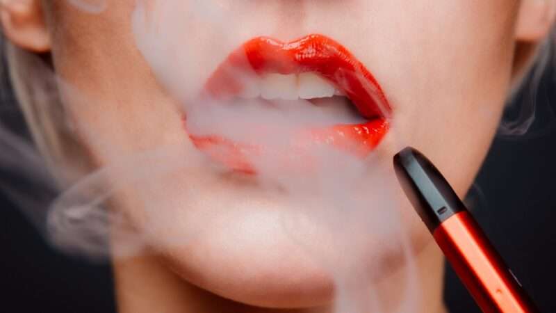 Woman with red lipstick has vapor coming out of her open mouth, with a vape pen near her lips.