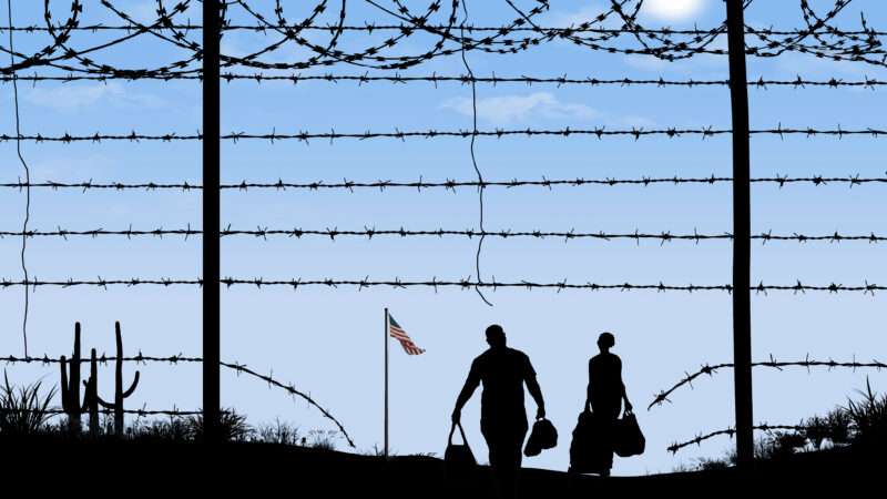 shadowed figures of two people holding bags walk toward a flying American flag as seen through a hole in a barbed wire fence